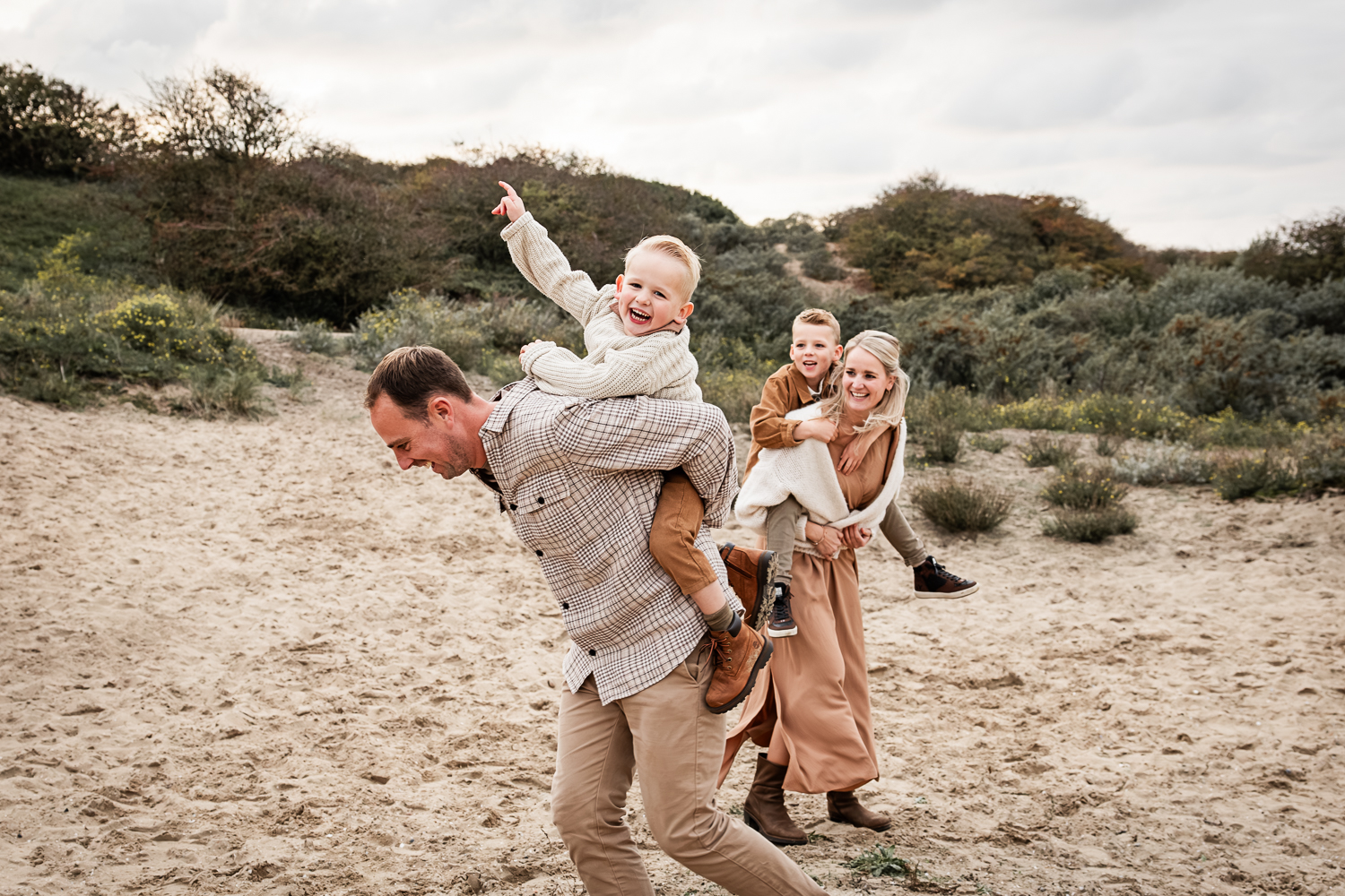 Twee kinderen zitten bij hun ouders op de rug en maken veel plezier tijdens een gezinsshoot in de duinen.